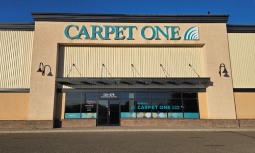 Front of Kimball Carpet One flooring store with the sign “CARPET ONE” in teal above an open entrance and glass windows, with small decorative finials on the columns under a clear blue sky.